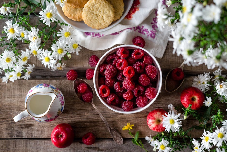 A rustic breakfast scene featuring a bowl of raspberries, apples, cookies, and white flowers on a wooden table.
