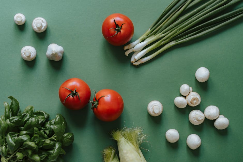Flat lay of fresh vegetables including tomatoes, mushrooms, and spring onions on a green surface.