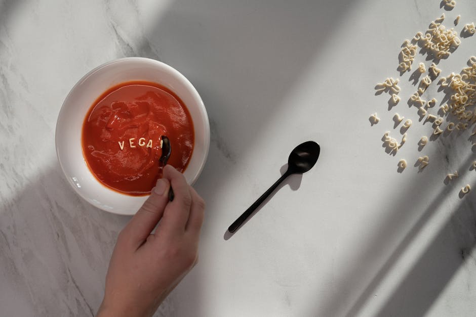 Flat lay of vegan tomato soup with alphabet pasta spelling 'VEGAN' on marble surface.