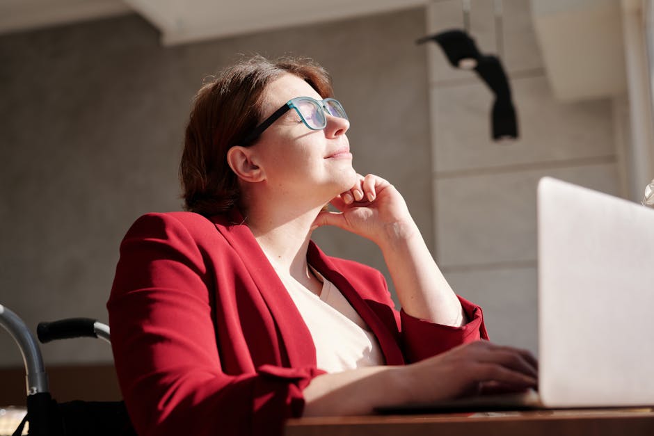 Woman in wheelchair wearing red suit and glasses, working on a laptop in a bright café.